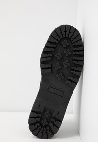 Semelle d'une botte noire avec des crampons profonds et des motifs rectangulaires et en forme de croix, positionnée à la verticale contre un fond d'angle blanc.