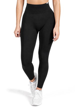 Frau trägt schwarze Leggings mit hohem Bund und weiße Sneakers, steht auf einem Bein mit entspannten Armen seitlich vor weißem Hintergrund.