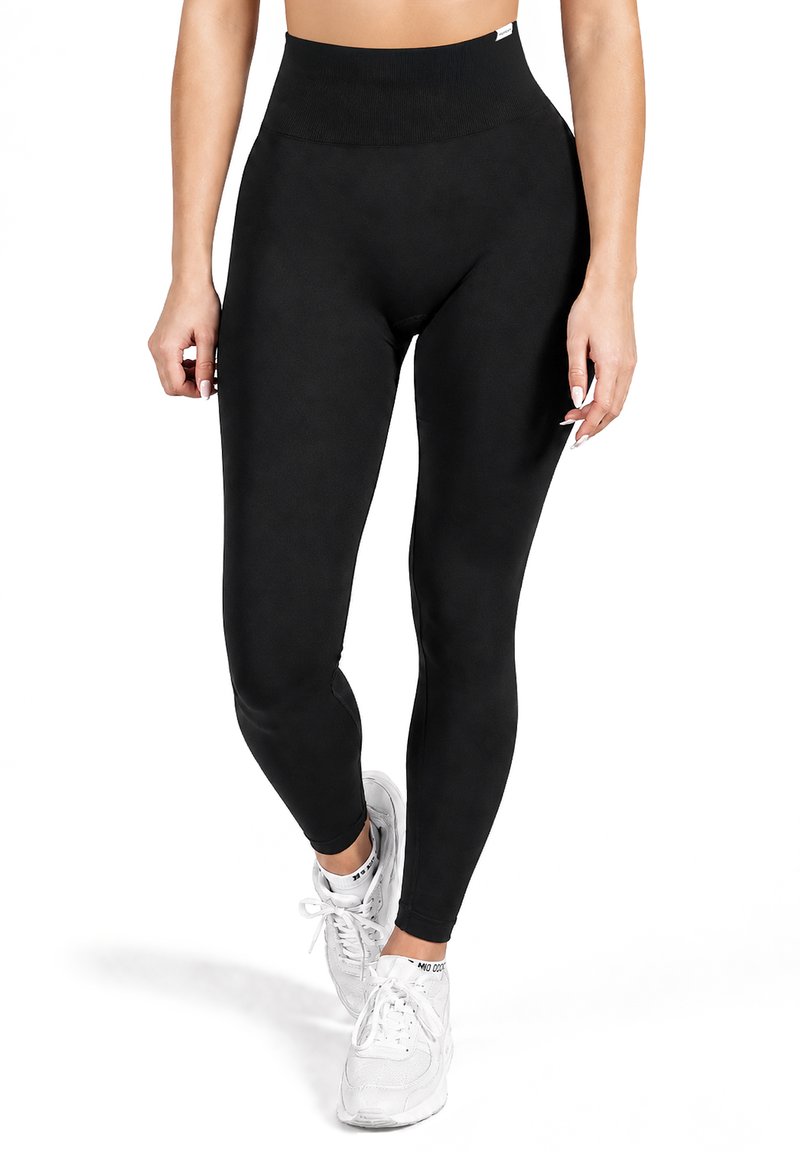 Femme portant un legging noir taille haute et des baskets blanches, debout sur un pied, les bras détendus le long du corps, sur fond blanc.