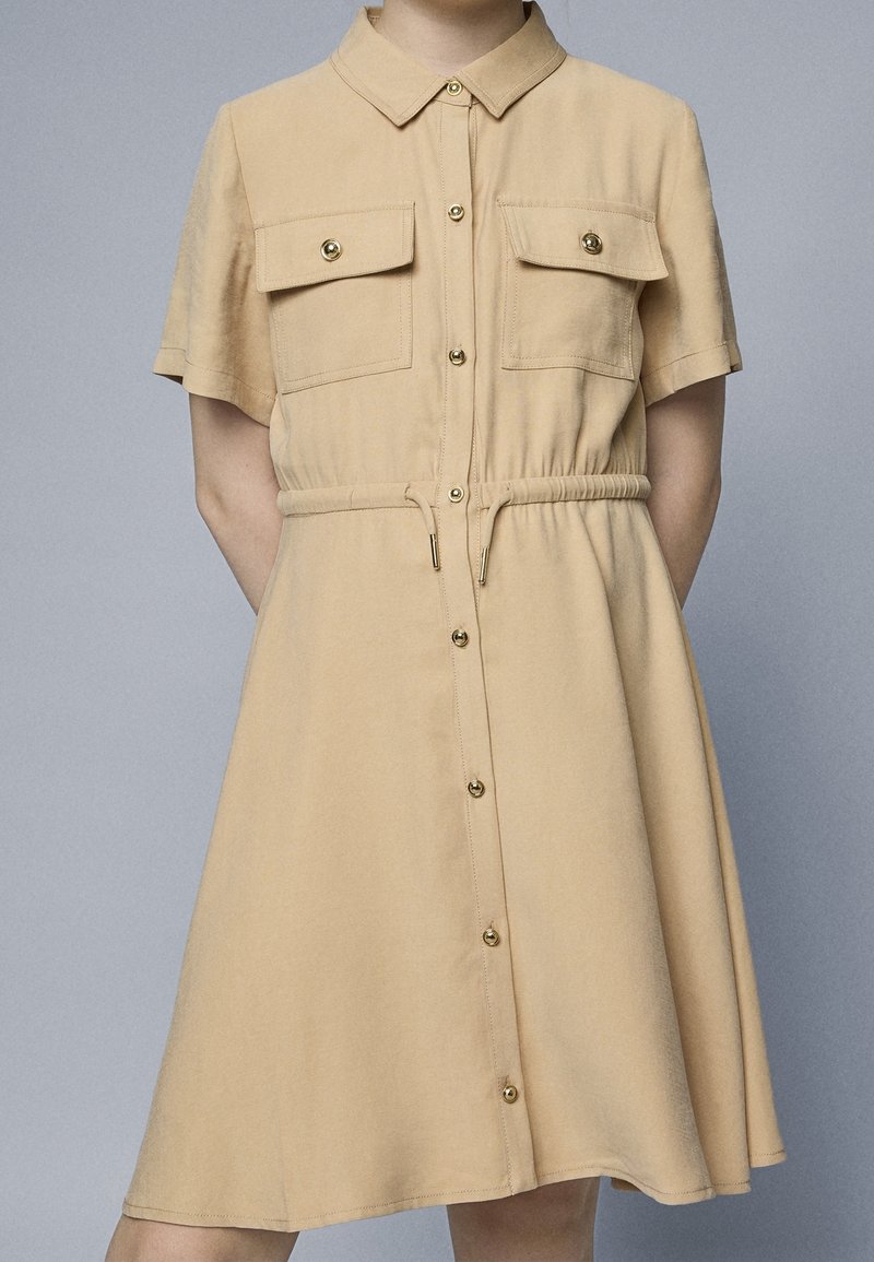 Robe chemise beige à manches courtes avec devant boutonné, deux poches poitrine à rabat, col et cordon de serrage à la taille portée par une personne.