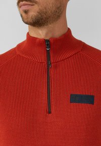 Rode ritssluitingtrui met een geribbelde textuur, hoge kraag en een marineblauw logo-vignet op de borst. Heeft een zwarte ritssluiting.