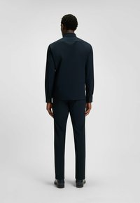 Camicia blu navy su misura con colletto, maniche lunghe e polsini con bottoni, abbinata a pantaloni su misura coordinati e scarpe nere. Tessuto dalla superficie liscia.
