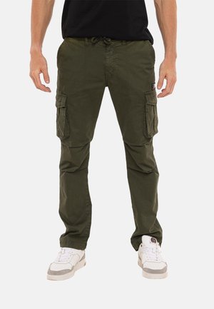 Homme portant un pantalon cargo vert olive avec des poches latérales et des baskets blanches, se tenant contre un fond blanc.