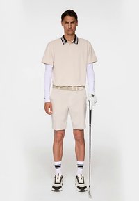 Beige pikétröja med svart randig krage, kombinerad med beige shorts. Vita långärmade tröjor under pikétröjan, beige bälte, vit golfhandske och skor.