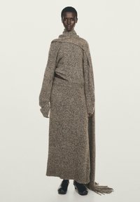 Gebreide jurk in een neutrale mix van bruin en beige, met een hoge hals, lange mouwen en een franjesaccent dat over één schouder hangt.