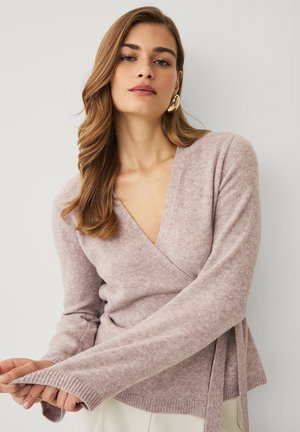 Kvinde iført en blød lyserød sweater med lange ærmer og gyldne hoop-øreringe, poserer mod en ensfarvet lys baggrund.