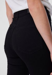 Detaljni prikaz osobe koja nosi crne traperice visokog struka s stražnjim džepovima i bijelu majicu uvučenu unutra.