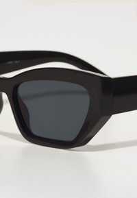 Ochelari de soare negri cu un design geometric, lentile pigmentate și brațe groase, drepte. Material neted cu un finisaj lucios.