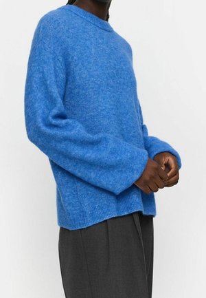 Persoon draagt een losse, gestructureerde blauwe trui en donkergrijze broek, staand tegen een effen lichte achtergrond met handen losjes gevouwen.