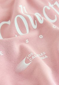 Světle růžová bavlněná tkanina s bílým textem a logem. Textura je hladká s mírným pružením a design obsahuje výrazné grafické prvky.