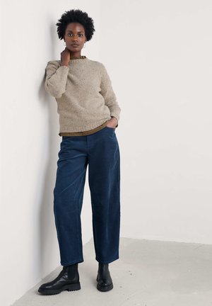 Pulover tricotat bej cu guler côt, asortat cu pantaloni largi din corduroy bleu închis și botine negre. Design simplu, texturat.
