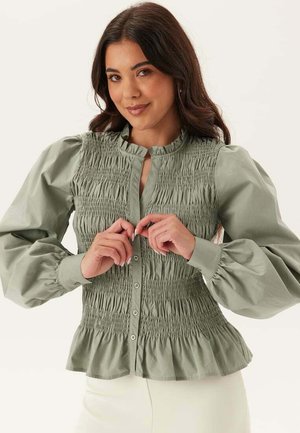Kvinde med langt mørkt hår iført en lysegrøn smocket bluse med puffede lange ærmer, knappet foran og med flæsekant, der knapper.