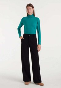 Jersey de cuello alto en color verde azulado, con mangas largas, combinado con pantalones de pierna ancha de talle alto en negro. La modelo está de pie con una mano en el bolsillo, frente a un fondo neutro.