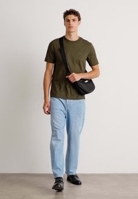 T-shirt vert foncé, jeans bleu clair, sac bandoulière noir, chaussures noires ; modèle se tenant contre un fond blanc uni.