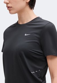 Άτομο που φοράει μαύρη αθλητική μπλούζα Nike με κοντό μανίκι και λευκό λογότυπο στο στήθος, φαίνεται από τους ώμους μέχρι πάνω από τη μέση.
