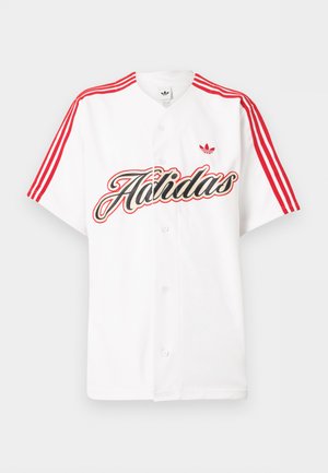 Chemise blanche à manches courtes avec des rayures rouges Adidas sur les épaules, un grand texte stylisé "Adidas" et un logo trèfle rouge sur la poitrine.
