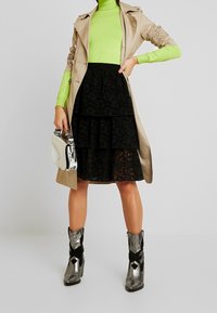 Manteau trench beige, haut à manches longues vert, jupe en dentelle noire à plusieurs niveaux, bottes de style cowboy métalliques argent et noires, et un petit sac à main brillant.