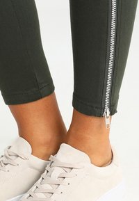 Enkel-lange donkergroene broek met een zilveren rits aan de zijkant, gedragen met lichtbeige suède veterschoenen.