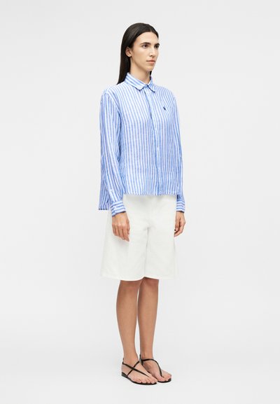 Femme debout portant une chemise à manches longues à rayures verticales bleues et blanches, un short blanc jusqu'aux genoux et des sandales plates noires.