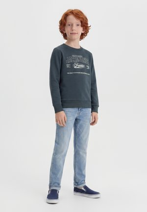 Garçon aux cheveux roux debout portant un sweat-shirt noir Jack & Jones, un jean bleu clair et des chaussures slip-on bleu marine sur un fond blanc uni.