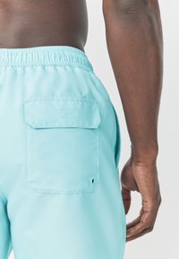 Turkooise zwemshorts met een elastische tailleband, een achterzak en een knoopdetail. De stof lijkt glad en lichtgewicht.