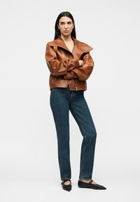 Woman with long dark hair wearing a brown leather jacket, blue jeans, and black pointed flats, standing with arms crossed.