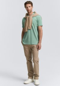 Grøn polo-shirt med korte ærmer, beige sweater draperet over skuldrene, beige slim-fit bukser og hvide sneakers. Simpelt, rent design.