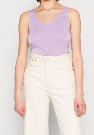 Kobieta w bezrękawniku w kolorze lawendy, wsuniętym w wysokowalne, kremowo-białe spodnie z widocznymi guzikami i kieszeniami.