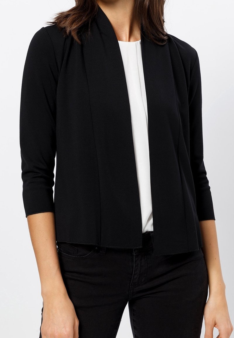Femme portant un cardigan noir ouvert sur le devant avec des manches trois-quarts, par-dessus un haut blanc et un pantalon noir sur un fond uni.