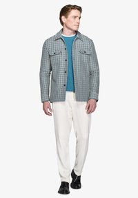 Flerfarvet jakke i blå, hvid og sort stof over en teal strikket sweater, parret med cremefarvede bukser og sorte sko.