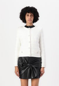 Veste blanche texturée à manches longues, col rond et boutons sur le devant, associée à une jupe noire en simili cuir avec un design noué.