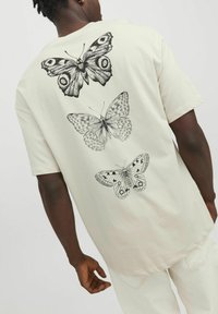 Light beige t-shirt featuring three detailed black butterfly prints on the back, with varying wing patterns and sizes. Short sleeves, cotton material.