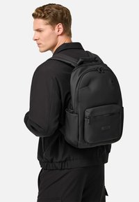 Mochila negra de nylon suave con un diseño redondeado, que cuenta con un bolsillo frontal con cremallera, correas ajustables y un asa superior.