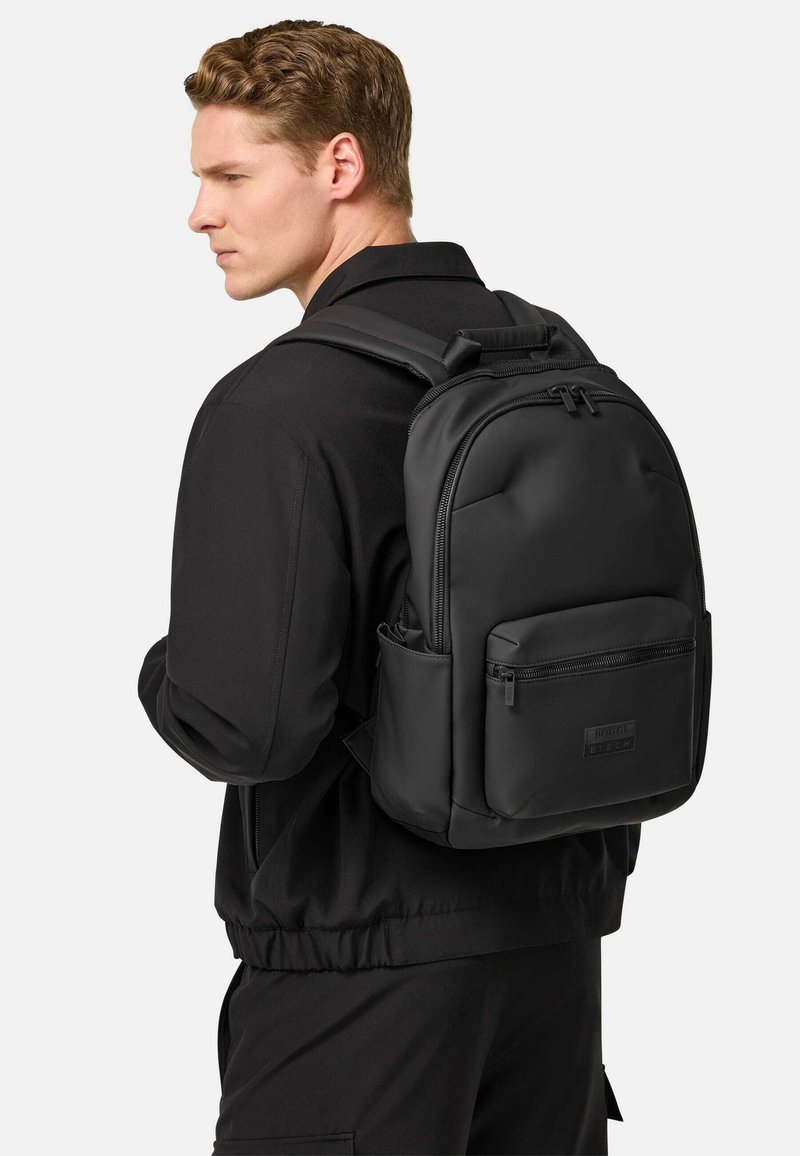 Mochila negra de nylon suave con un diseño redondeado, que cuenta con un bolsillo frontal con cremallera, correas ajustables y un asa superior.