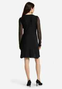 Schwarzes Kleid mit transparenten, puffigen langen Ärmeln und einem taillierten Oberteil. Der Rock ist knielang und mit verstreuten Pailletten verziert, was einen strukturierten Look erzeugt.