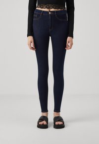 Jeans skinny a vita alta blu scuro con cuciture arancioni, tasche frontali e chiusura con bottone, abbinati a sandali neri.