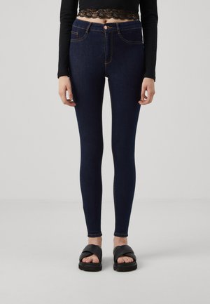 Jeans skinny a vita alta blu scuro con cuciture arancioni, tasche frontali e chiusura con bottone, abbinati a sandali neri.