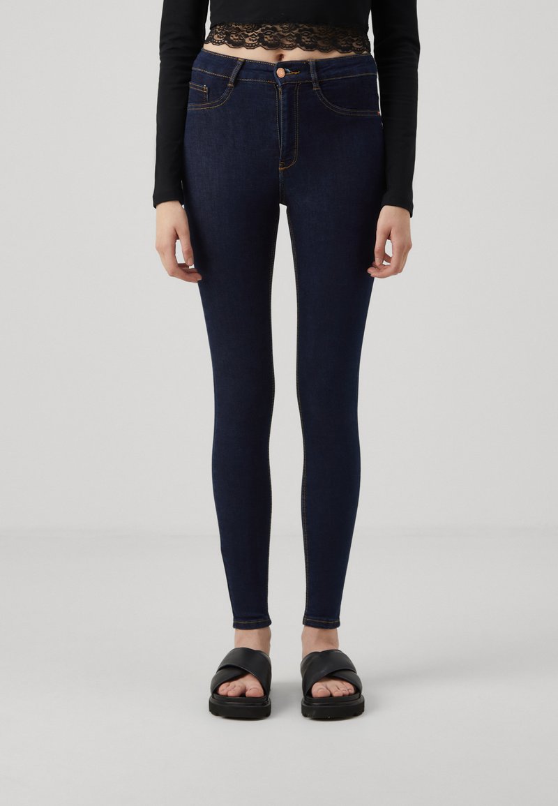 Jeans skinny a vita alta blu scuro con cuciture arancioni, tasche frontali e chiusura con bottone, abbinati a sandali neri.