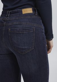 Tmavě modré džíny z denimu s viditelnou koženou nášivkou označenou "fransa DENIM." Obsahují standardní zadní kapsy s jemnými švy.
