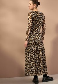 Robe maxi à imprimé léopard avec des manches longues, présentant une jupe plissée, un tissu transparent et un décolleté subtil. Des chaussures noires à semelle épaisse complètent le look.