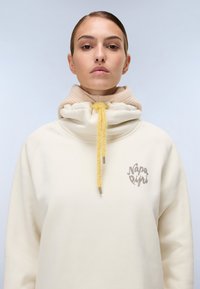Kremowy sweatshirt z wysokim kołnierzem, z beżowym wewnętrznym kołnierzem. Żółdy sznurek do regulacji i akcent z logo z przodu. Miękka tekstura materiału.