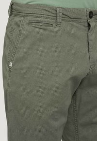 Pantalons en coton vert texturé avec des motifs en losange, dotés d'une fermeture à bouton et de poches latérales. Gros plan sur le tissu et les d étails de couture.