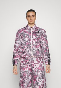 Giacca cropped e pantaloni coordinati in un motivo astratto rosa e grigio. Presenta chiusura con zip, colletto e strisce laterali nere. Tessuto morbido.