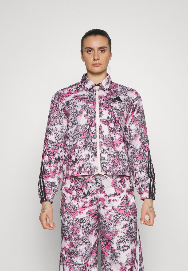 Giacca cropped e pantaloni coordinati in un motivo astratto rosa e grigio. Presenta chiusura con zip, colletto e strisce laterali nere. Tessuto morbido.