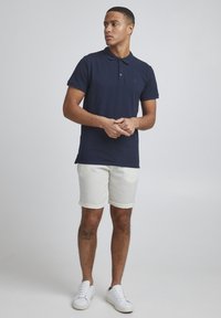 Camiseta polo azul marino con mangas cortas y cuello, combinada con pantalones cortos de mezclilla de color claro. Presenta zapatillas blancas y un tatuaje visible en la pierna.