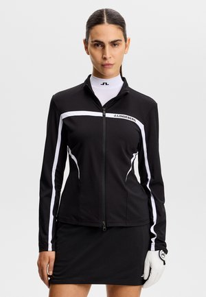 Vrouw die een zwart jack met ritssluiting en witte strepen draagt, een zwarte rok en een witte golfhandschoen aan haar linkerhand, staand tegen een effen achtergrond.