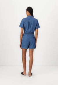 Denim overal s krátkým rukávem, límečkem a elastickým pasem. Materiál je středně modrý denim s ohrnutými kraťasy.