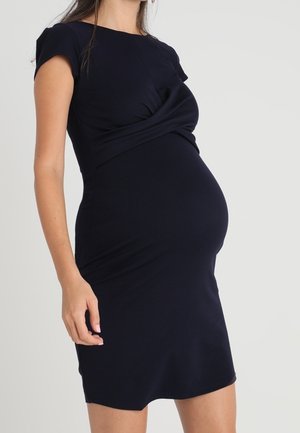 Mujer embarazada vistiendo un vestido negro ajustado hasta la rodilla con mangas cortas, sobre un fondo liso y claro.