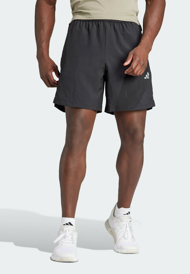 Homme portant un short de sport noir, des chaussures de sport blanches et des chaussettes blanches, debout devant un fond clair uni.