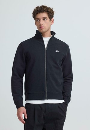 Giovane uomo con capelli ricci che indossa una giacca nera con cerniera sopra una maglietta bianca, in piedi contro uno sfondo chiaro uniforme.